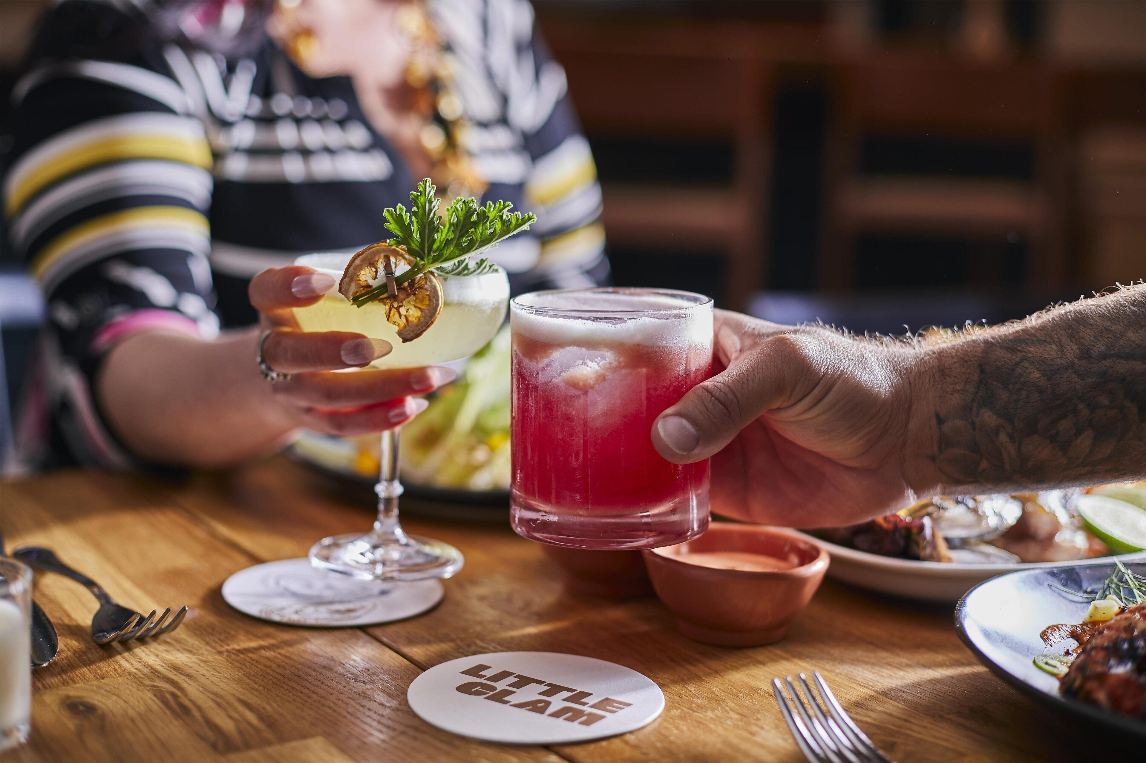 A person holding a glass with a red drink, another glass with a yellow drink, and a plate with food.