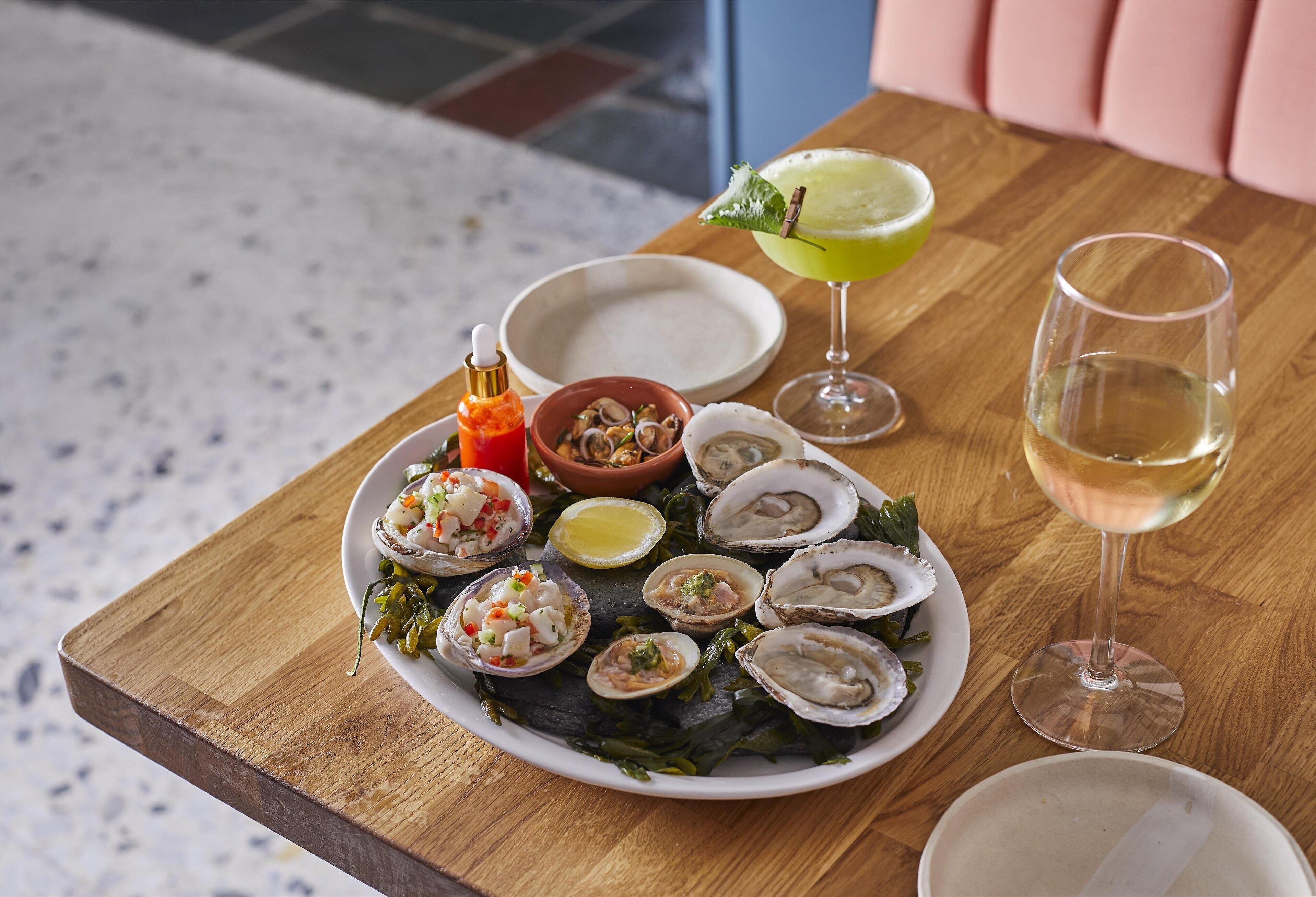 Una mesa de madera con un plato de mariscos, un cóctel y una copa de vino blanco.