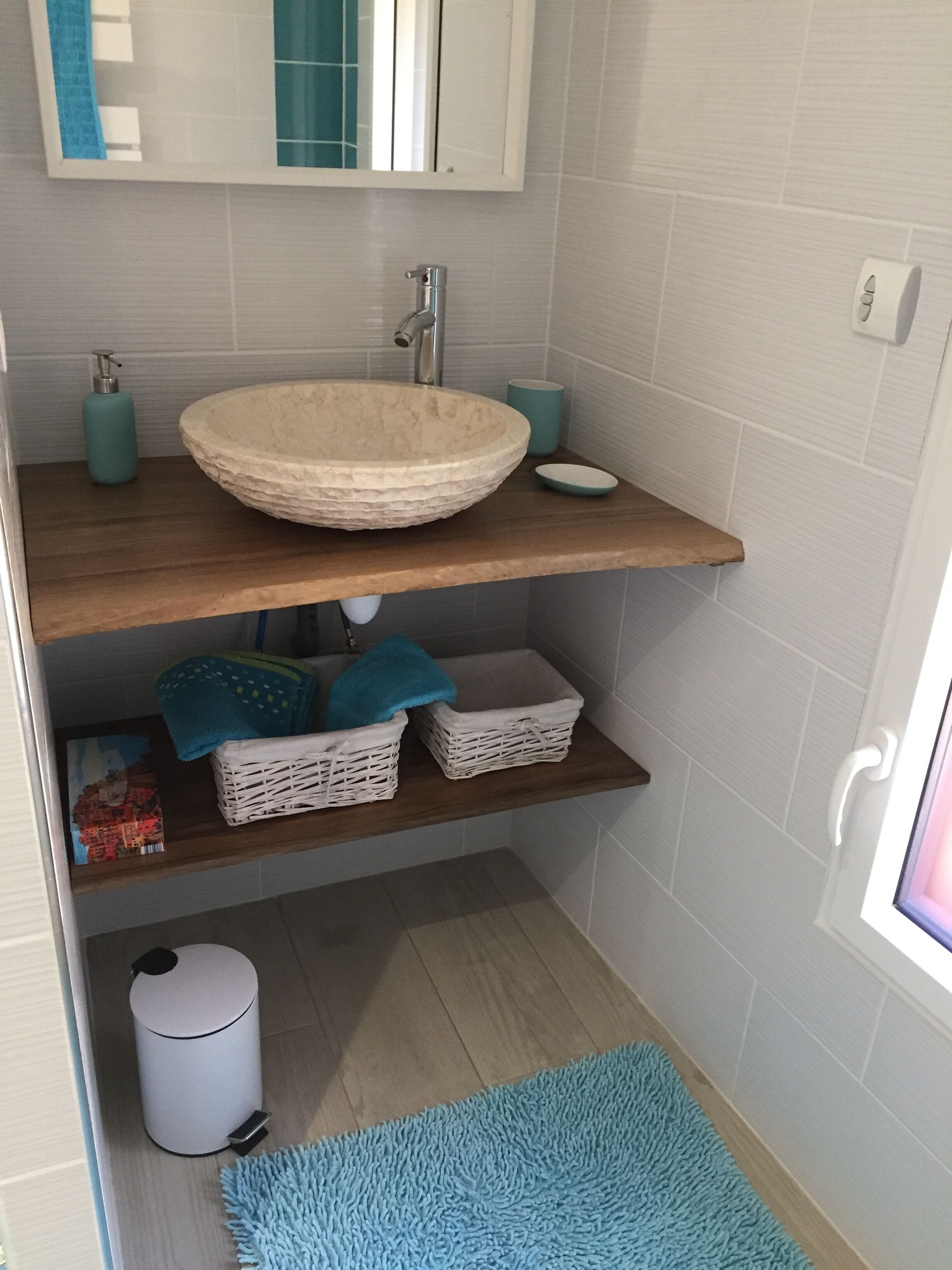 A bathroom with a round basin, a mirror, a shelf with baskets, a trash can, and a blue rug.