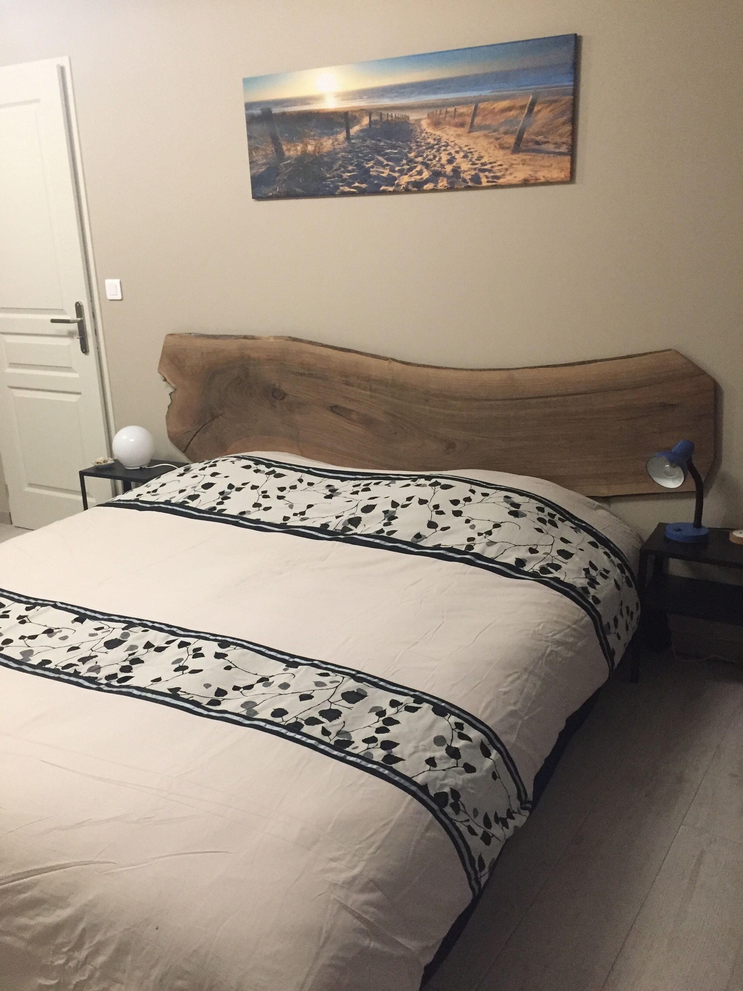A bedroom with a wooden headboard, a bed with a floral-patterned bedspread, a nightstand with a lamp, and a painting of a beach scene on the wall.