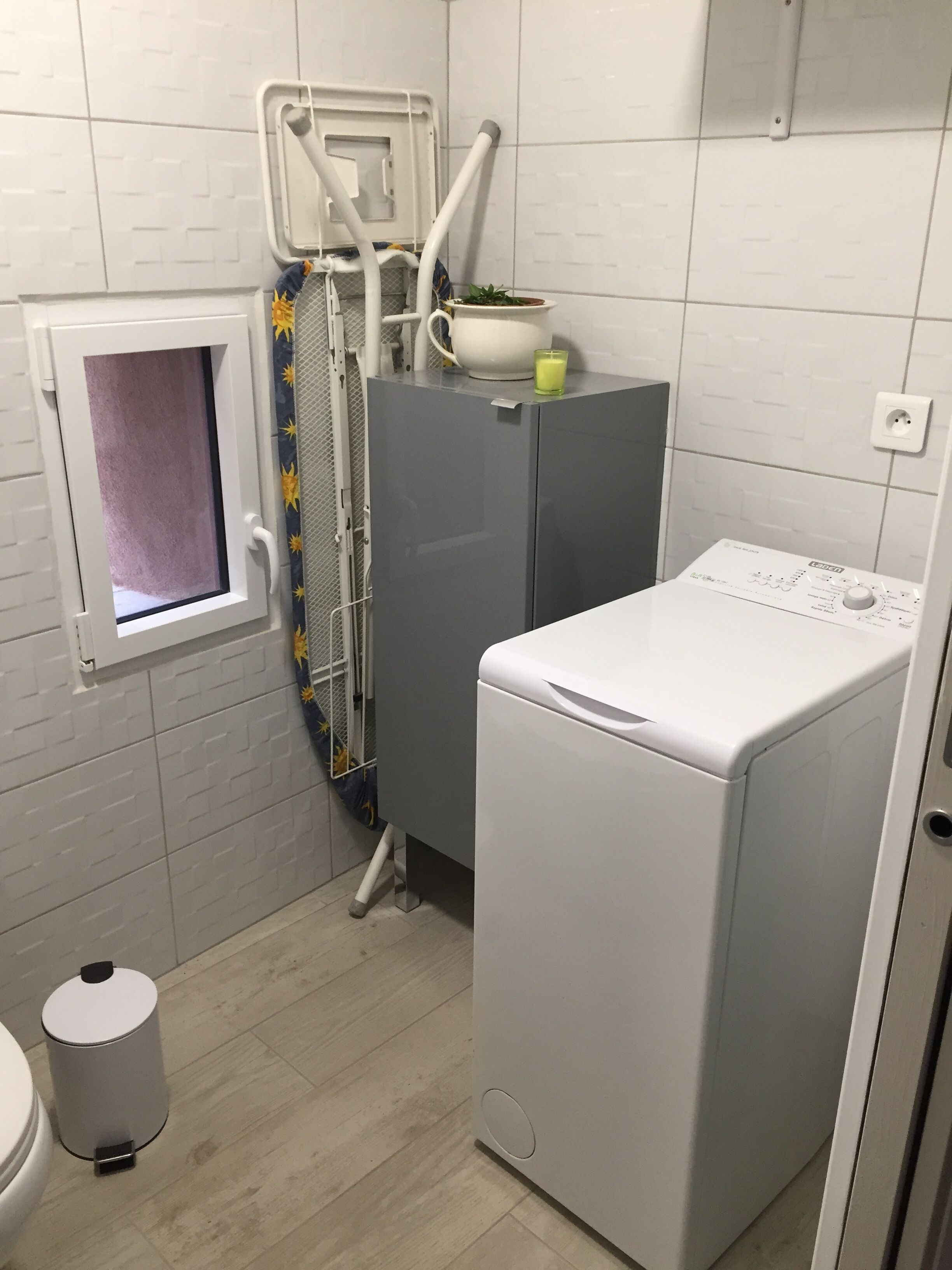 Un baño pequeño con lavadora, una ventana con cortina, un cubo de basura y un calentador de pared.