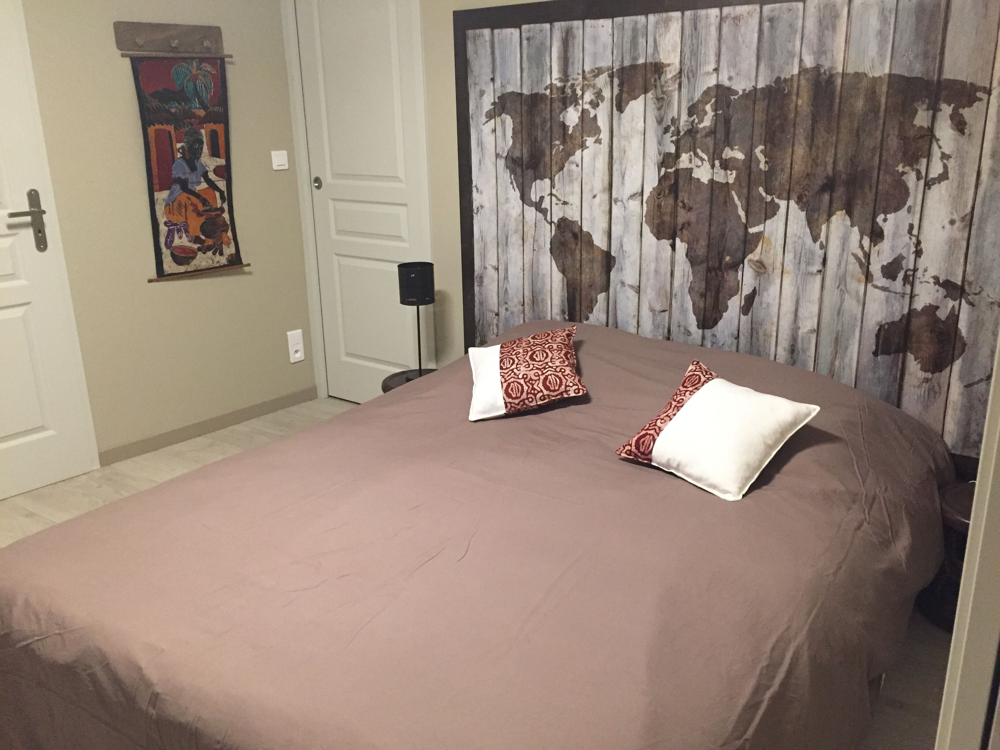 A bedroom with a large bed, two patterned pillows, a world map wall, a door, and a small painting on the wall.