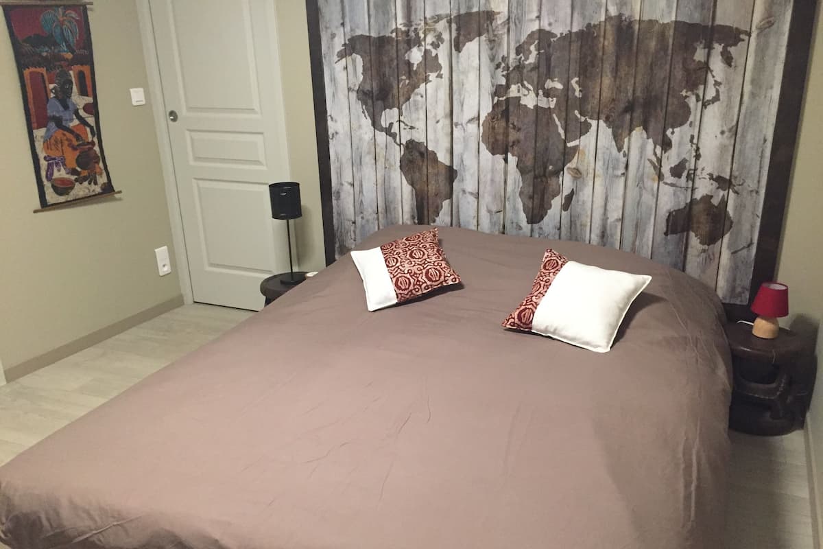 A bedroom with a large bed, two pillows, a world map curtain, a small table with a lamp, and a door.