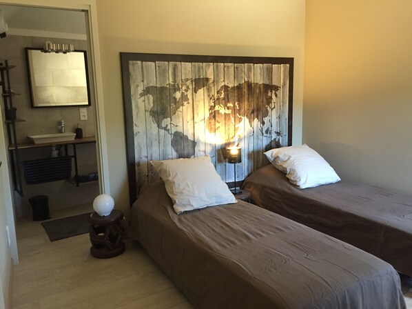 Une chambre avec deux lits, une tête de lit en bois ornée d’une carte du monde, une petite table avec une lampe, et une salle de bain visible à travers une porte ouverte.