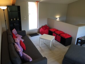 Un salon moderne avec un canapé gris foncé, une table basse blanche et des fauteuils en forme de poire rouge. On peut également apercevoir une bibliothèque noire et une petite table avec des magazines.