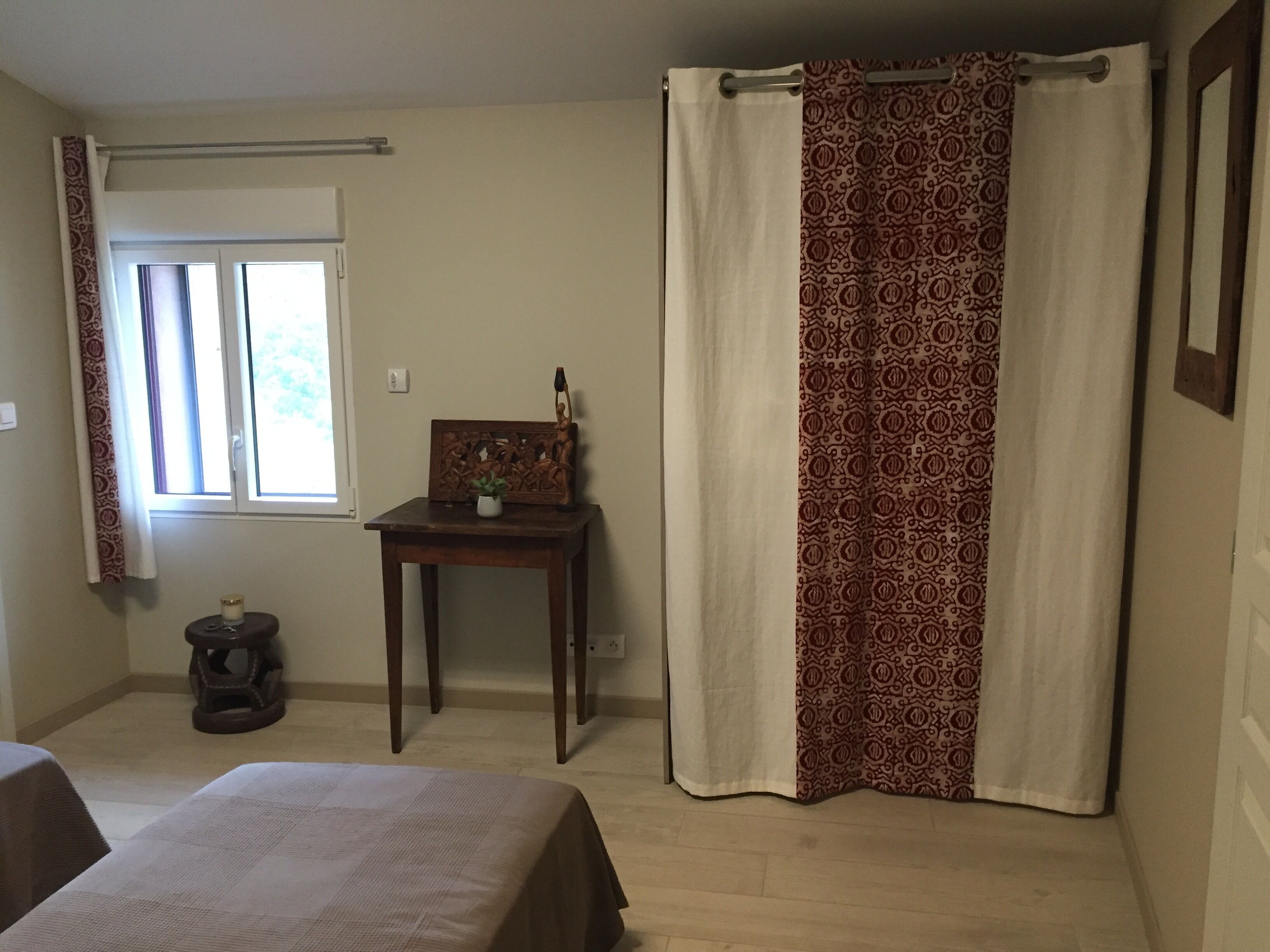 Une chambre à coucher avec un lit, une petite table sur laquelle est posée une plante, une fenêtre avec des rideaux et un rideau de douche à motifs.