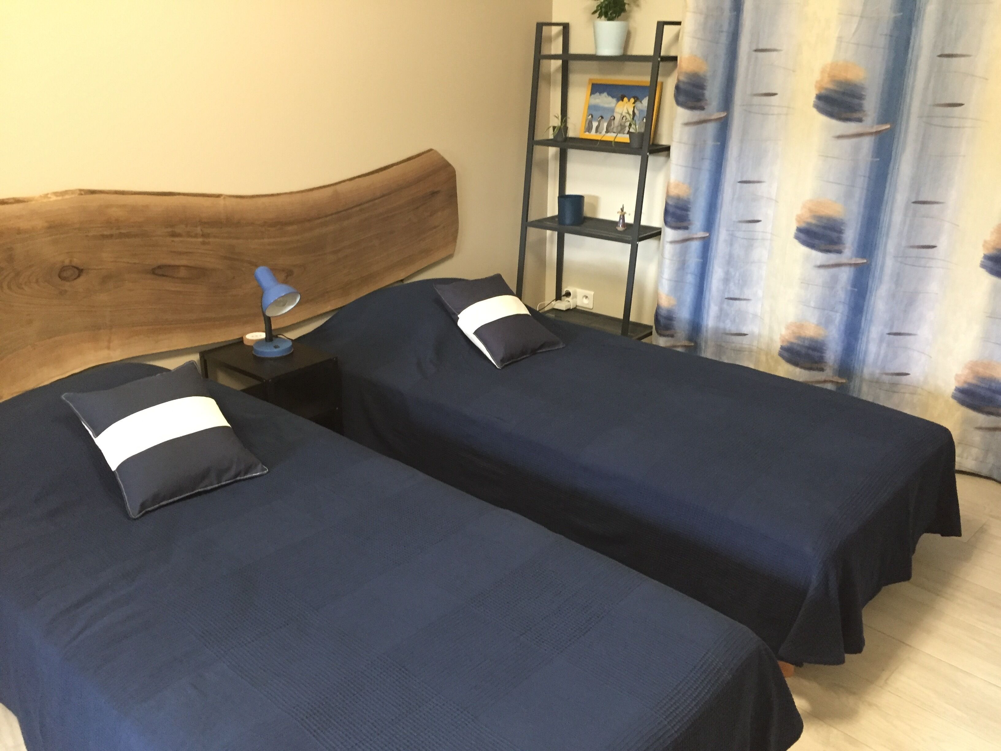 Una habitación con dos camas individuales, cabecera de madera, mesita de noche con lámpara, estante con objetos decorativos y cortina estampada con hojas.