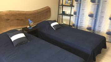 Una habitación con dos camas individuales, cabecera de madera, mesita de noche con lámpara, estante con objetos decorativos y cortina estampada con hojas.