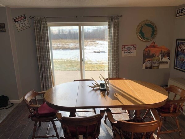 A dining area with a round table and chairs, a view through a glass door to a snowy outdoor area, and various wall decorations including a welcome sign and a Budweiser logo. - ATV and Snowmobile trails 1/4 mile away. Full size Big Buck Hunter Arcade game. (Danbury)