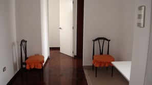 Um corredor com duas cadeiras de madeira, uma pequena mesa e uma porta branca no final, com um interruptor de luz na parede.