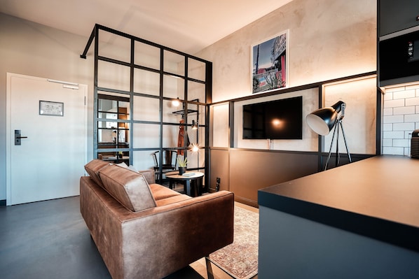 Una sala de estar moderna con un sofá de cuero marrón, una mesa de comedor con sillas, un televisor montado en la pared y una zona de cocina con mostrador y armarios.