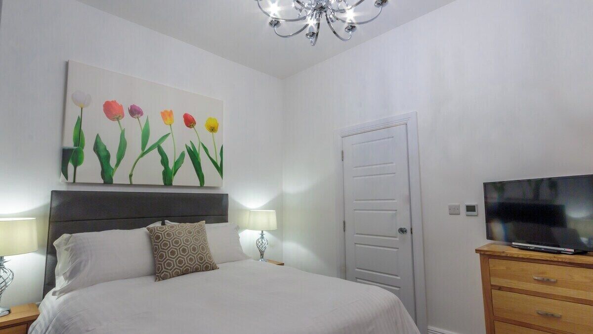 A bedroom with a bed, a TV on a wooden dresser, a painting of flowers on the wall, and a door.