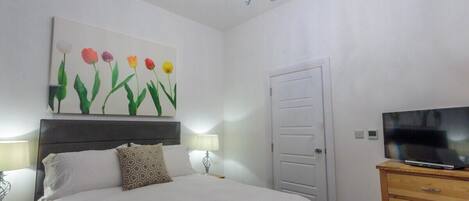 A bedroom with a bed, a TV on a wooden dresser, a painting of flowers on the wall, and a door.