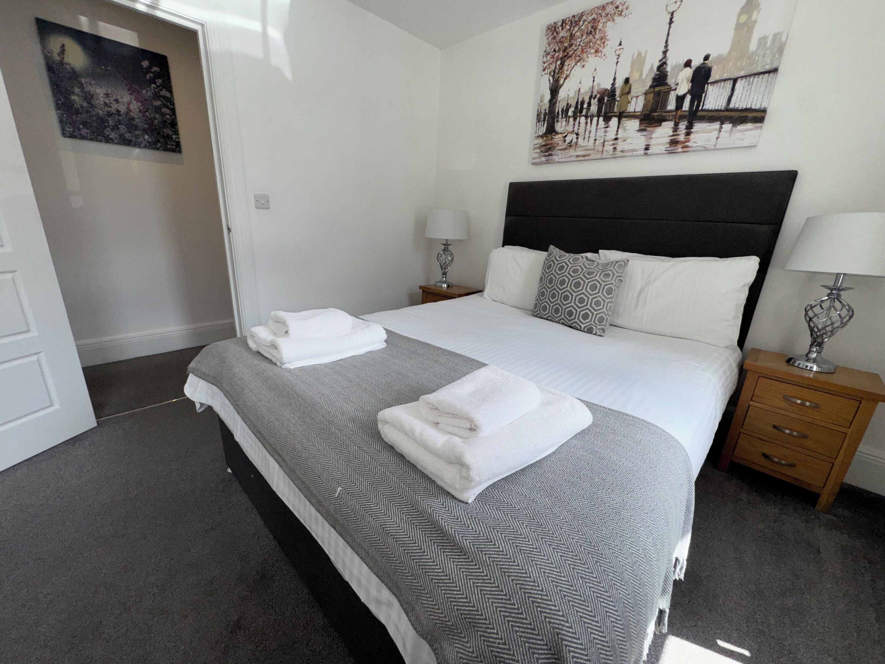 A neatly made double bed with a patterned bedspread, two bedside tables with lamps, a wall-mounted painting, and a door to the left.