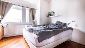 Une chambre à coucher comprenant un lit, une commode, un radiateur, une fenêtre avec des rideaux et une étagère ornée d’objets décoratifs.