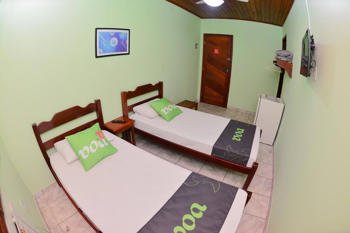 A room with two wooden beds, each with a green and white bedspread, a small bedside table, a wooden door, and a wall-mounted TV.