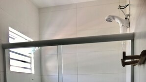 Una ducha a nivel con puerta de cristal, paredes con azulejos blancos, un cabezal de ducha y una ventana con persianas.