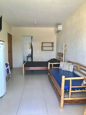 A room with two wooden beds, a small table, a wall-mounted shelf, and a refrigerator.