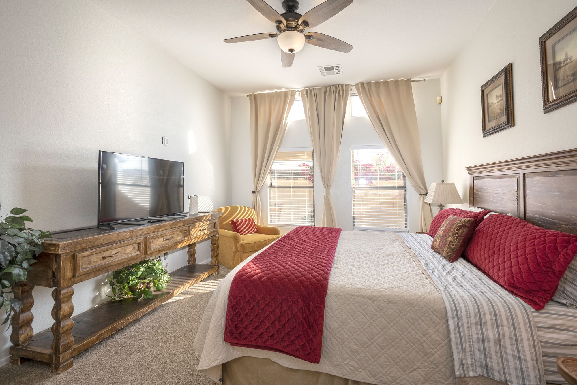 A bedroom with a bed, a TV on a wooden stand, a ceiling fan, and a window with curtains.