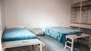 Une petite chambre propre, avec deux lits simples, une petite table, une armoire blanche et un lavabo blanc.