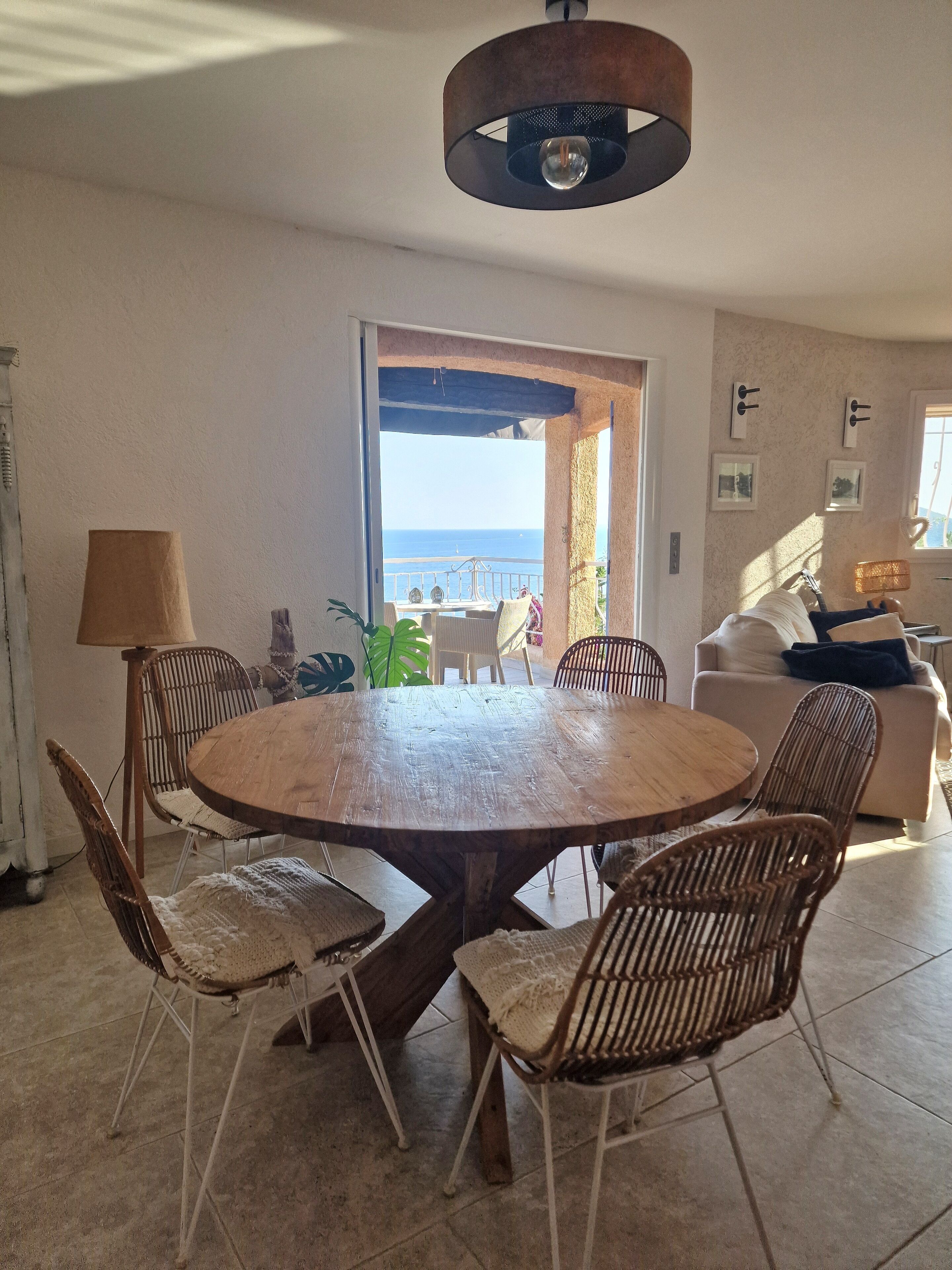 Een eetruimte met een ronde houten tafel en rieten stoelen, uitzicht op zee door een open deur en een balkon met tuinmeubilair.