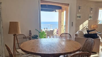 Een eetruimte met een ronde houten tafel en rieten stoelen, uitzicht op zee door een open deur en een balkon met tuinmeubilair.