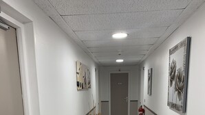 A clean, modern hallway with white walls, a wooden floor, and artwork on the walls. - Hotel bon accueil (Oyonnax)