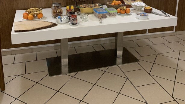 A buffet table with various food items and condiments, set against a wall with a wooden wreath and two wall-mounted lights. - Hotel bon accueil (Oyonnax)
