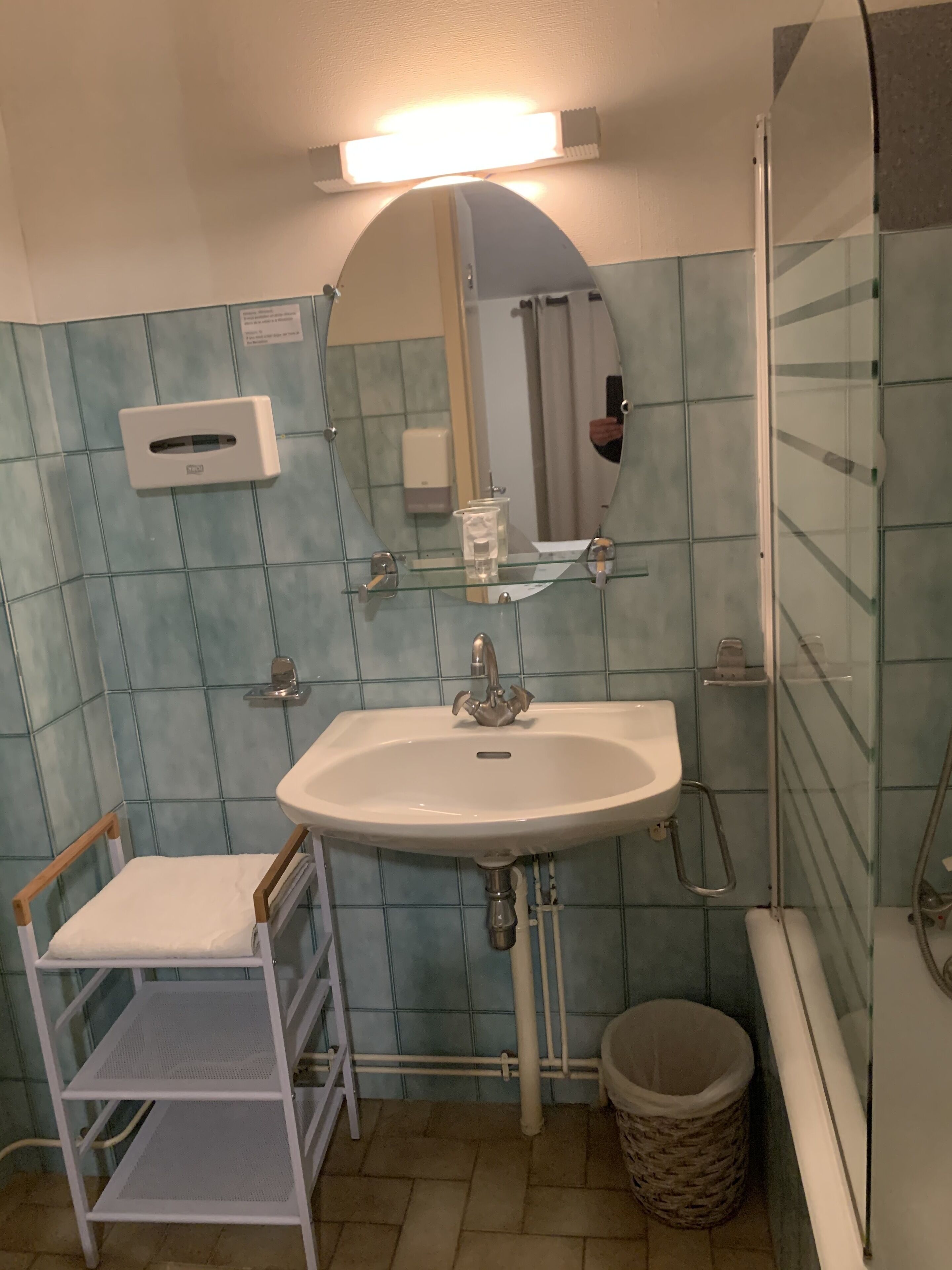 A bathroom with a white sink, a mirror, a glass shower enclosure, a hand dryer, and a small trash bin.