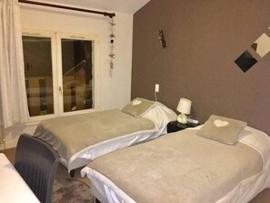 A room with two beds, a window with curtains, a bedside table with a lamp, and a wall decoration. - Hotel bon accueil (Oyonnax)