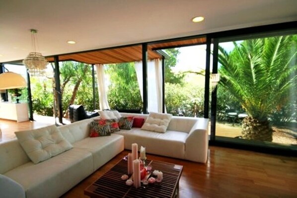 A modern living room with a white sofa, a coffee table with candles, and large windows offering a view of greenery.