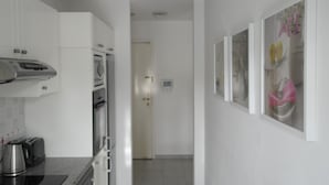A modern kitchen with white cabinets, a stainless steel range hood, a built-in oven, and a coffee maker. There is a hallway with a door leading to another room.