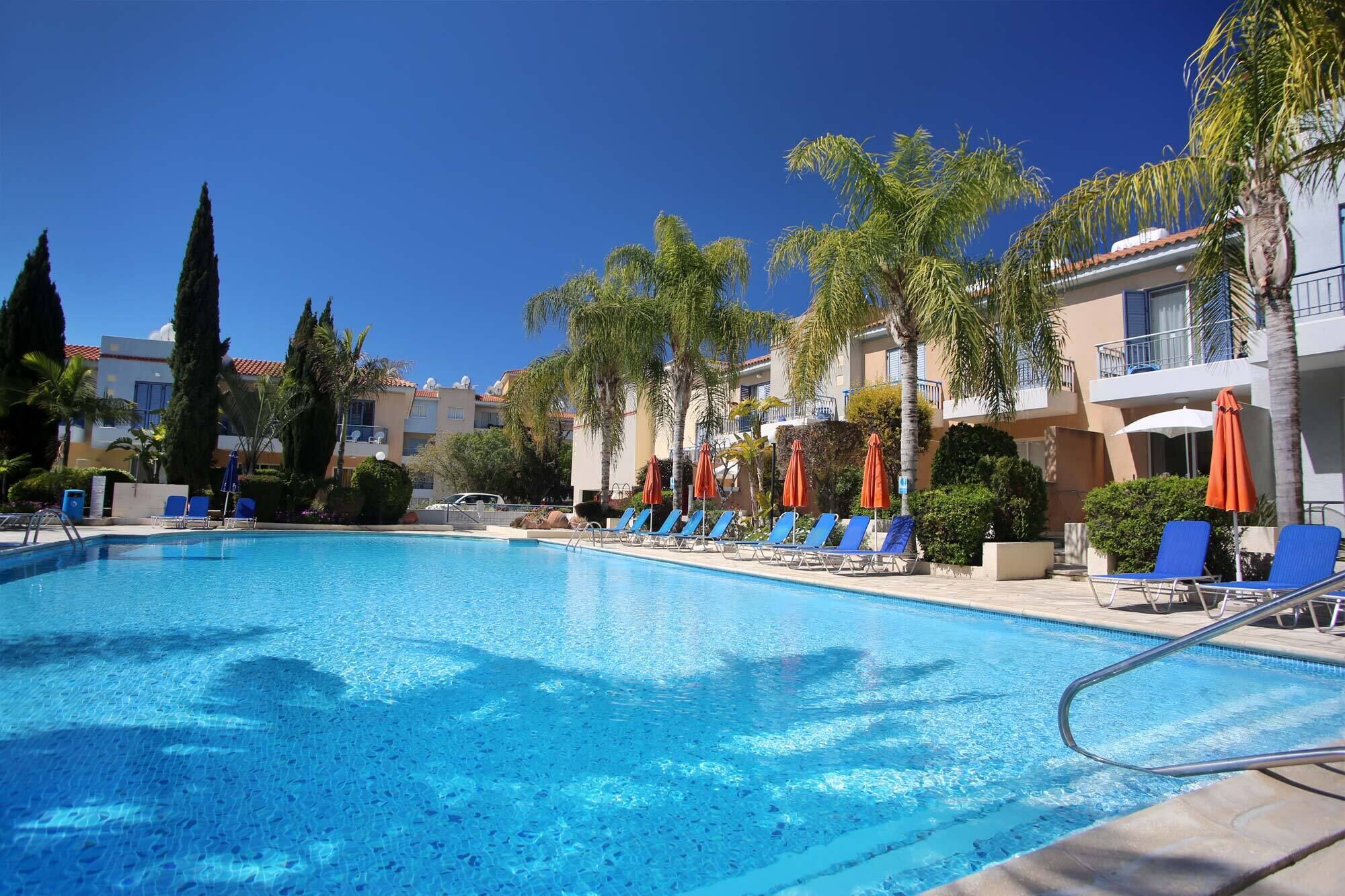 A swimming pool with lounge chairs and umbrellas, surrounded by palm trees and a multi-story building with balconies.