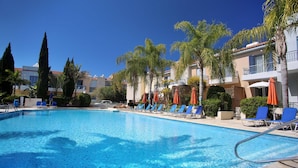 A swimming pool with lounge chairs and umbrellas, surrounded by palm trees and a multi-story building with balconies.