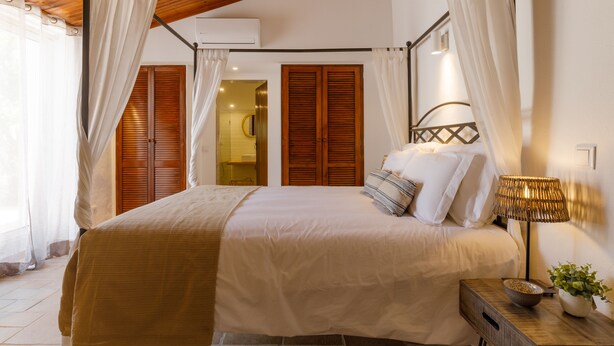 A bedroom with a bed, curtains, a bedside lamp, a plant, and a wooden headboard. - Monte da Luz - Casa da Mó (Luz)