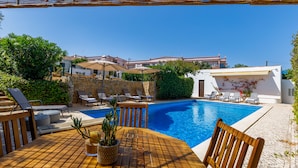 A poolside area with a wooden table, lounge chairs, and a stone wall. - Monte da Luz - Casa da Mó (Luz)