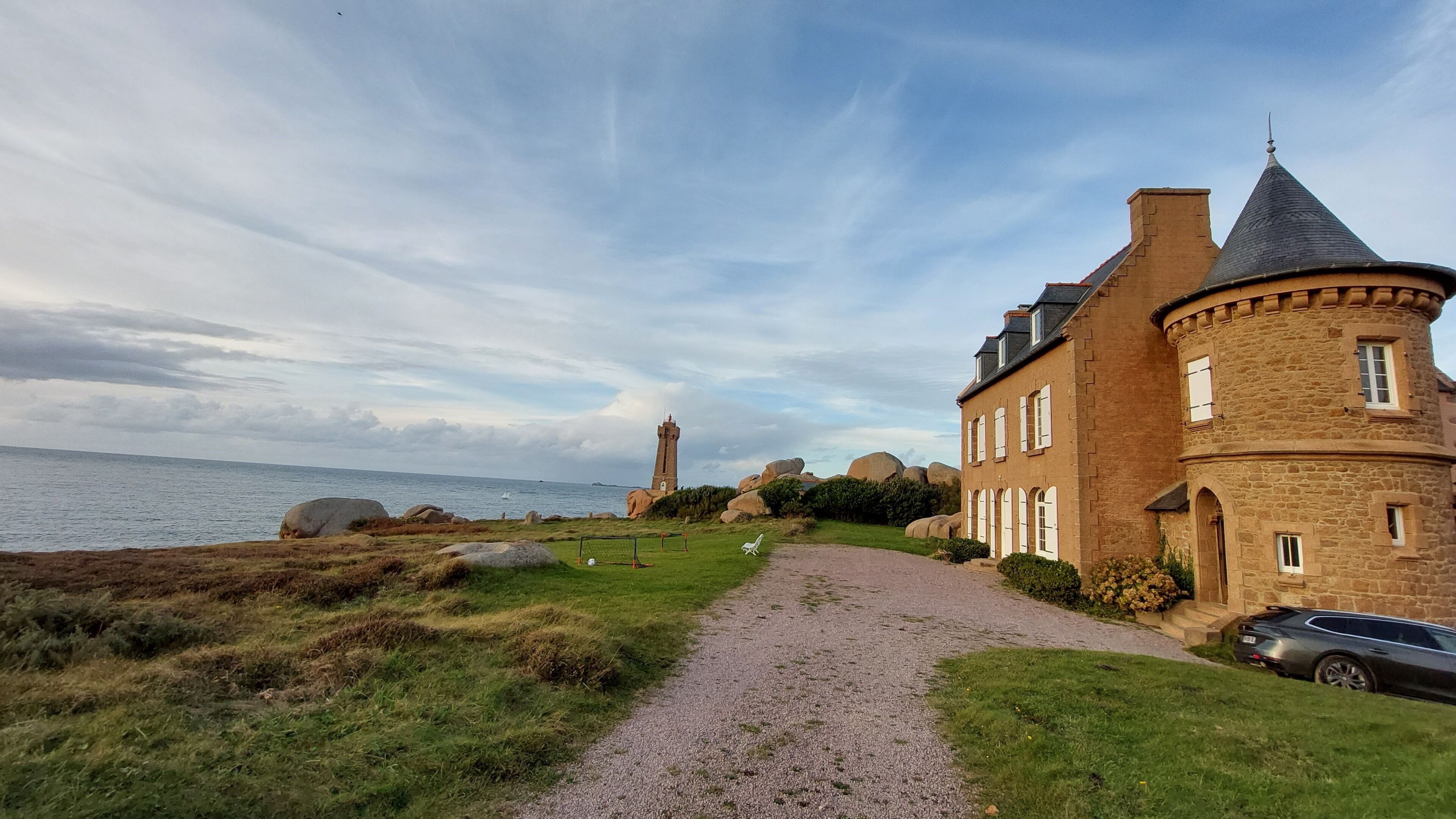 Ein Küstenhaus mit einem Kiesweg, der zum Meer führt, einem Leuchtturm in der Ferne und einem geparkten Auto in der Nähe.