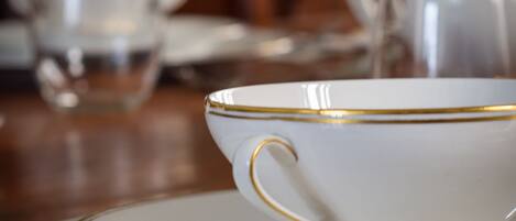 A white teacup with gold trim on a saucer, placed on a table with other teacups and a teapot in the background.