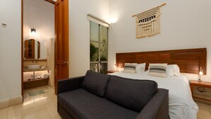 A hotel room with a bed, a sofa, a bedside table, a lamp, a wall-mounted macrame piece, and a bathroom with a sink and mirror. - Villa Orquidea Boutique Hotel (Mérida)