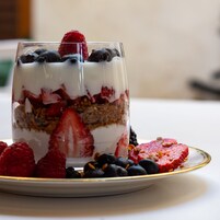 A layered dessert in a clear glass, garnished with berries and surrounded by more berries on a plate.