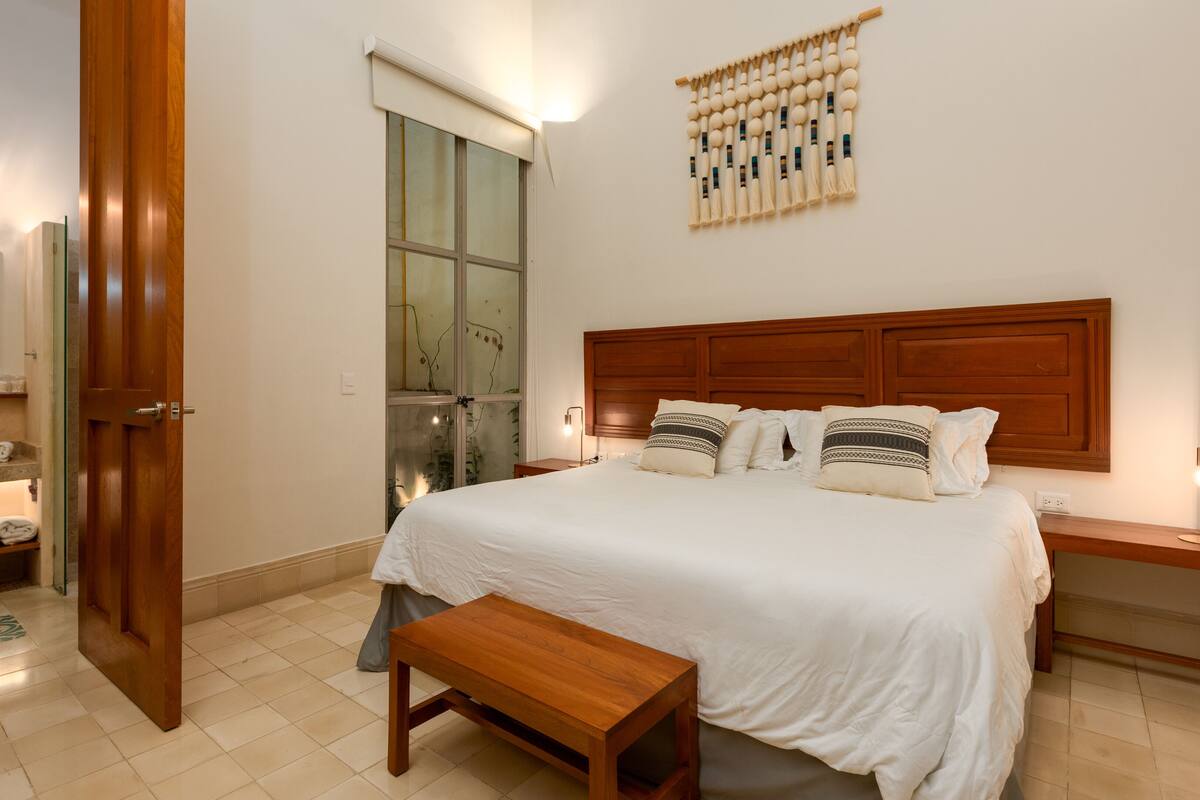 A bedroom with a wooden headboard, a large bed with white bedding, a bedside table with a lamp, and a wooden bench.