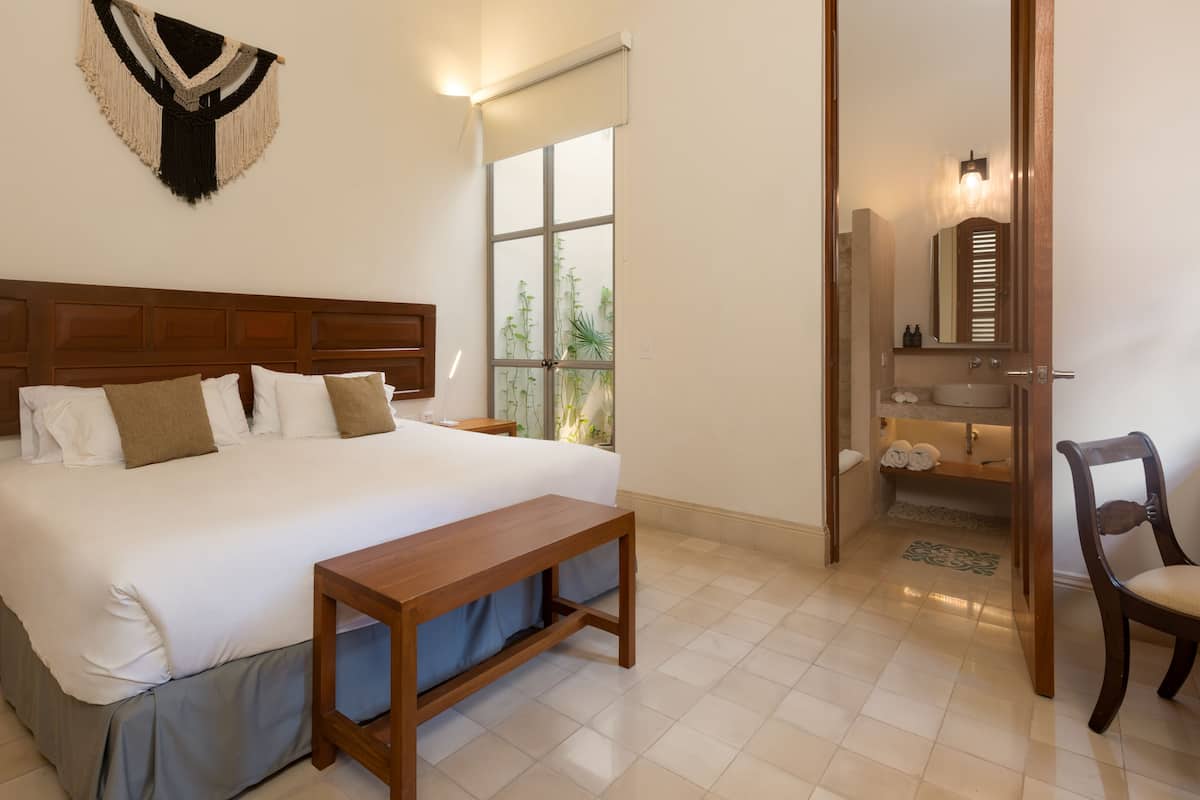 A neatly arranged hotel room with a large bed, bedside tables, a bench, a window with plants, and a bathroom with a sink and mirror.