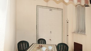Uma sala de jantar com uma mesa posta para dois, cadeiras, parede de azulejos, uma porta e uma janela com cortinas estampadas.