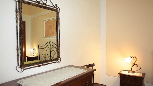 Quarto com cómoda de madeira, espelho com moldura elaborada e mesa de cabeceira com abajur.