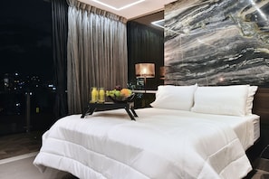 Een hotelkamer met een groot bed, een nachtkastje met een schaal fruit, uitzicht op de stad en een marmeren muur.