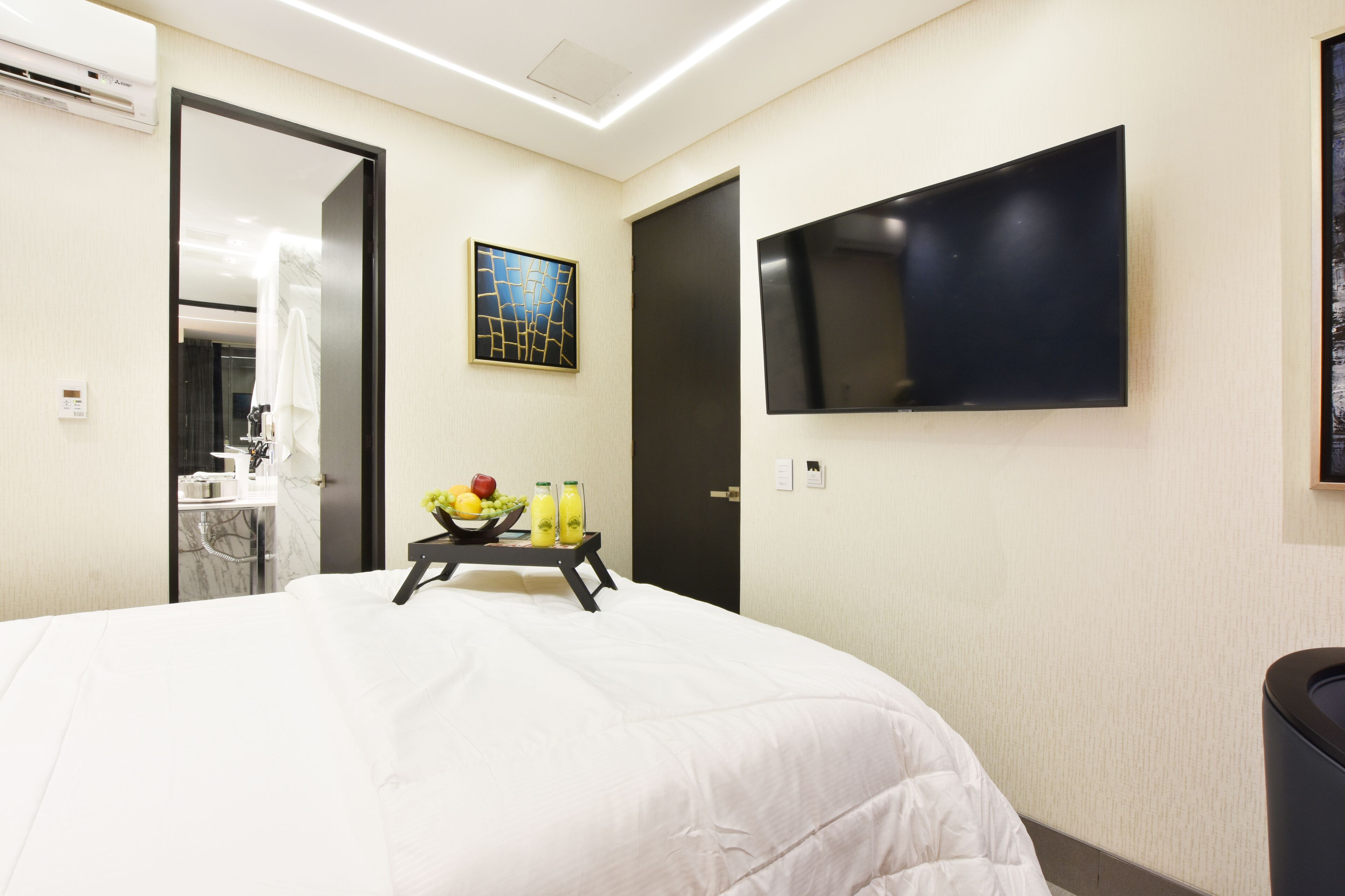 Ein Hotelzimmer mit einem großen Bett, einem flachbildfernseher an der Wand, einem kleinen Tisch mit Obst und Getränken sowie einem sichtbaren Badezimmer hinter einer offenen Tür.