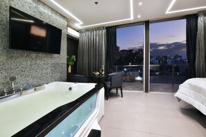 A modern hotel room with a large bathtub, a flat-screen TV mounted on a mosaic wall, a dining area with a table and chairs, and a balcony with a city view. - Luxury Combo Apartments with Jacuzzis and View (Medellín)