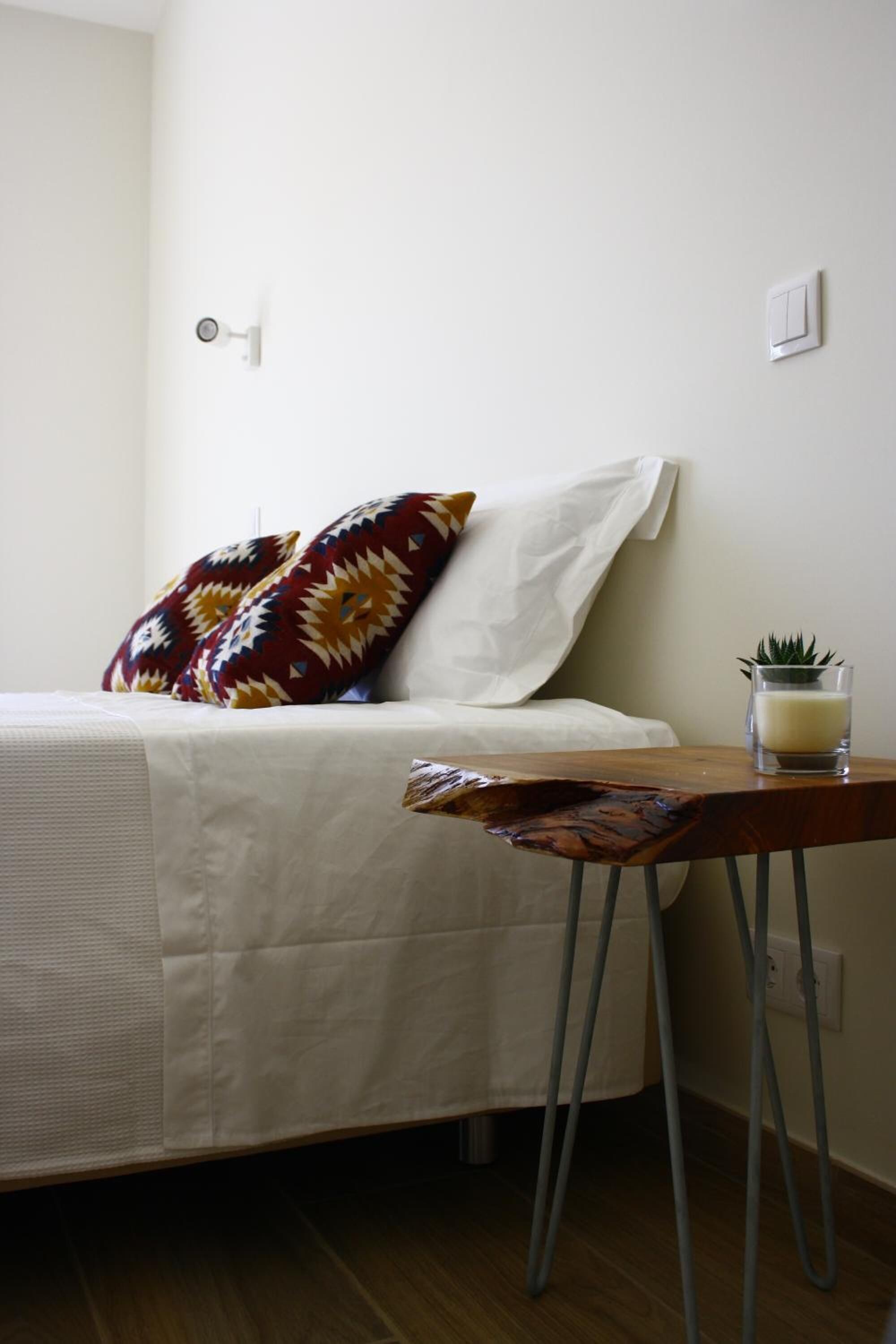 Un lit bien fait, avec une couette blanche et des coussins à motifs, une petite table sur laquelle se trouvent un verre et une bougie, et un interrupteur fixé au mur.