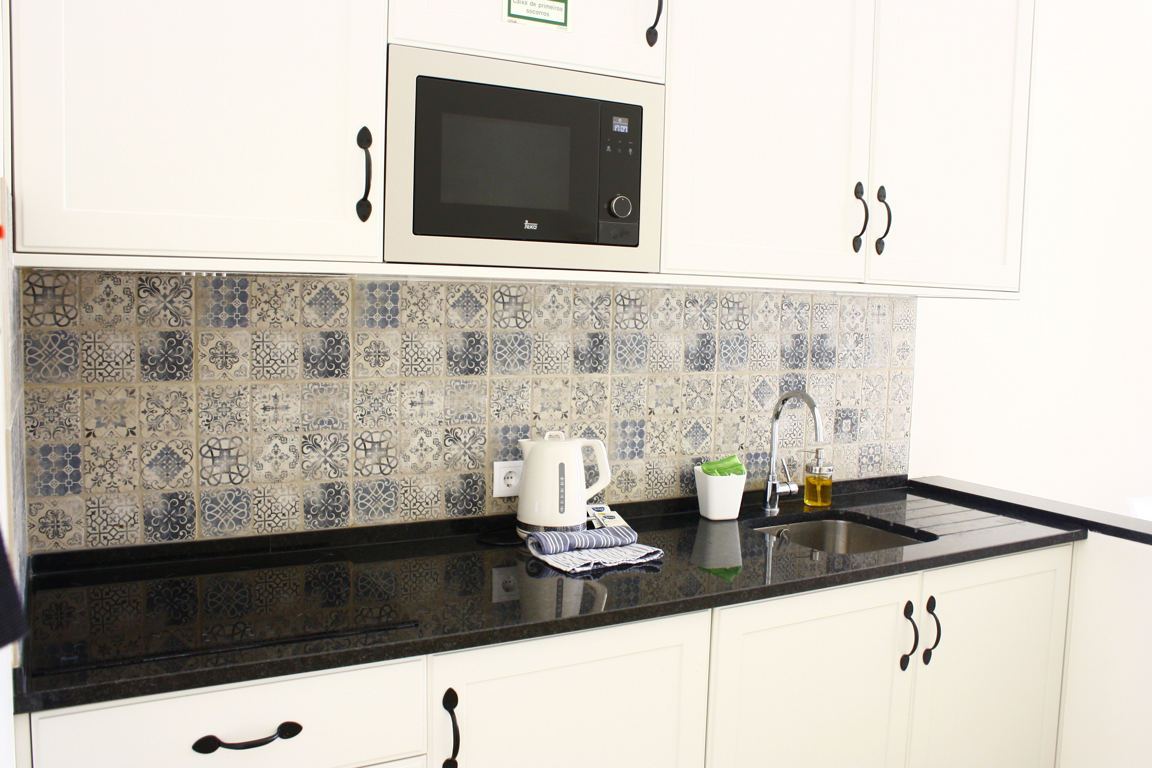 A kitchen with white cabinets, a black countertop, a microwave, a kettle, a coffee maker, and a sink.