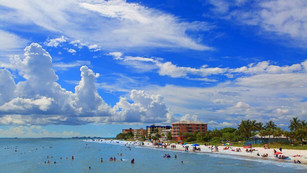 Una playa con cielo despejado, palmeras y un hotel al fondo.